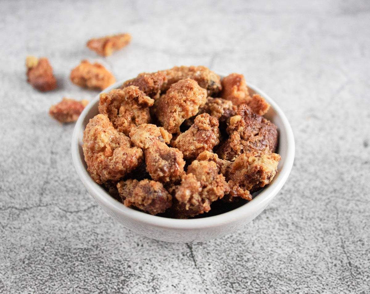 A bowl of delicious Mashuga nuts with a crusty, golden outer layer with only a few other nuts scattered in the background.